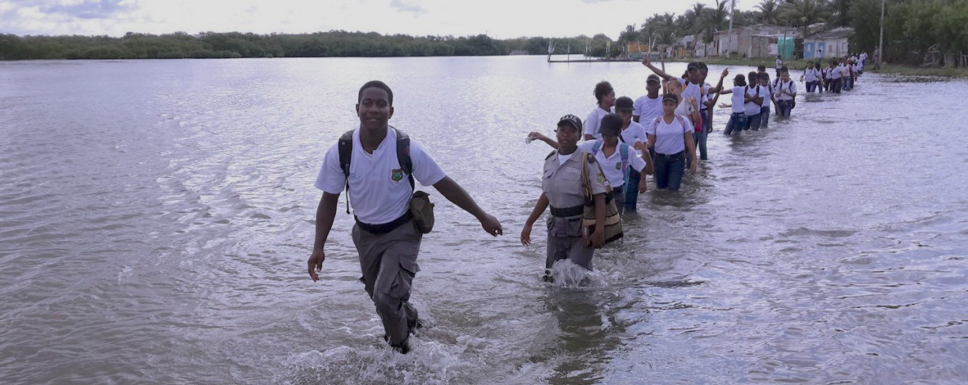 An image showing several youth in uniform in a single file line walking in ankle-deep water in what appears to be a lake or inlet.