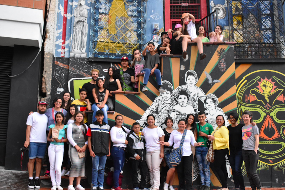 Group photograph of a large number of people posing in front of a colourfully painted building, some sitting on stairs and some standing in front