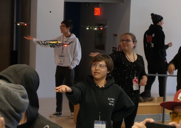 A video still showing young people gathered in a room, with their right arms outstretched and palms up, with drones hovering above their hands.