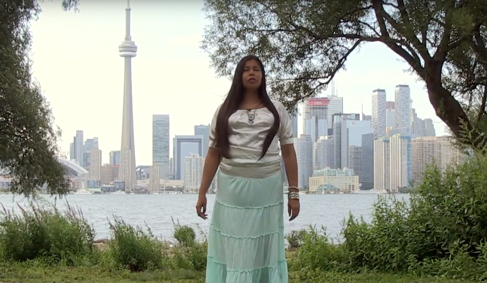 A photo showing an indigenous woman standing in front of some trees, grass and a lake, with the CN tower and Toronto skyline in the distance.